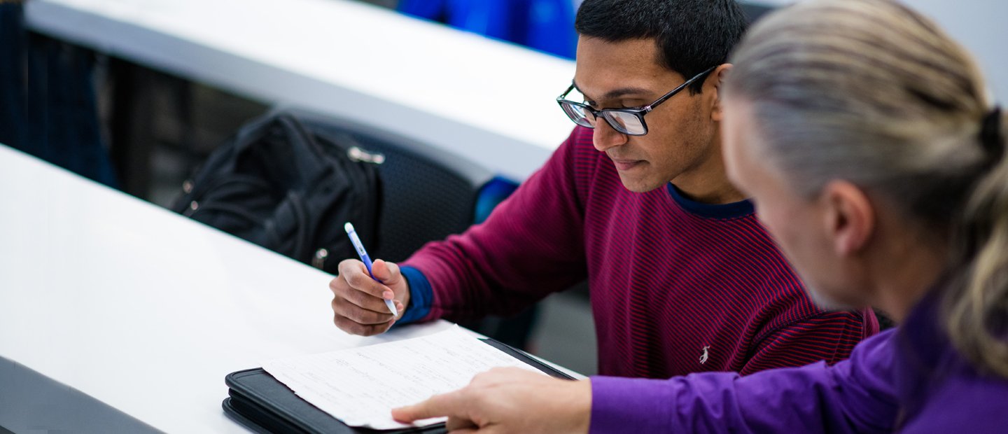 academics_adm Two men seated at a long white table, one holds a pencil while the other points to a piece of paper on the table