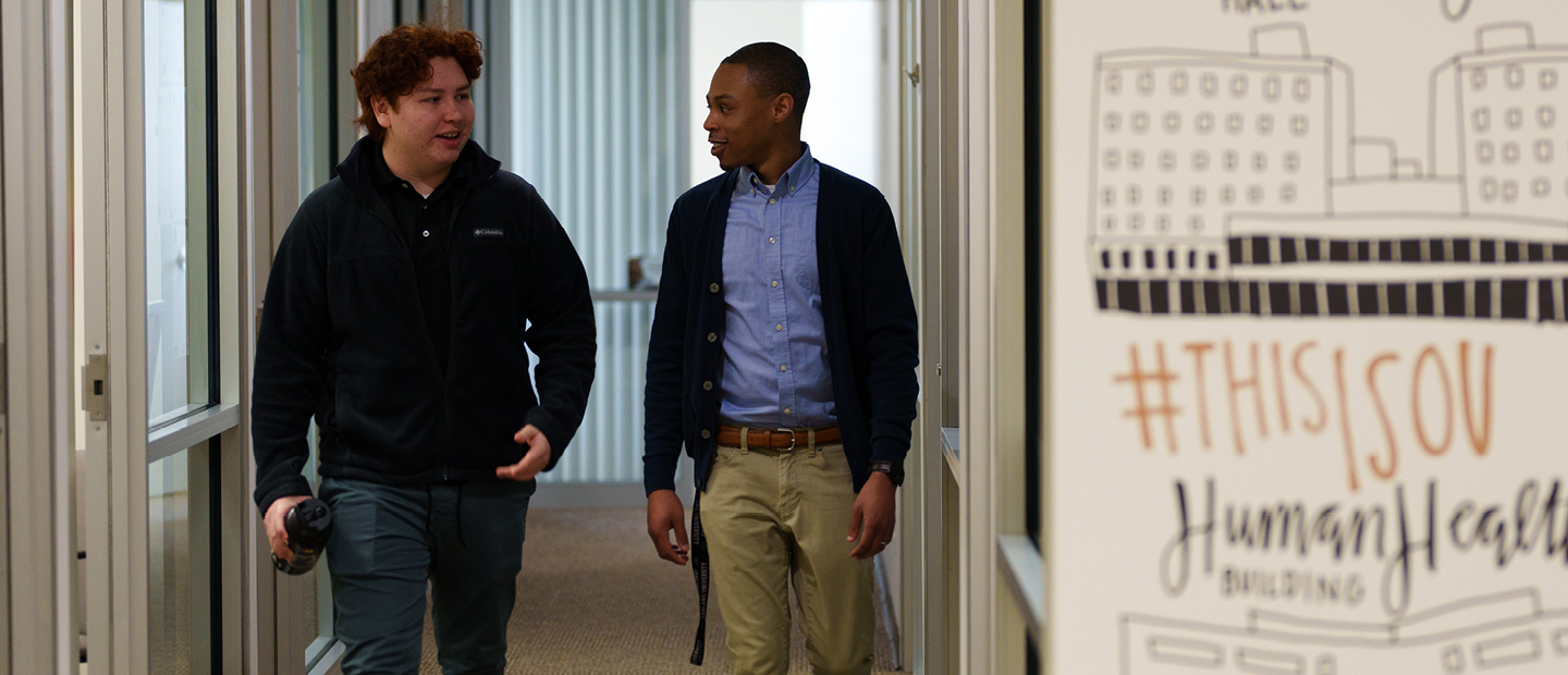 First Year Resources Adviser and student walking in the hallway, talking, with wall in foreground featuring drawings of campus features