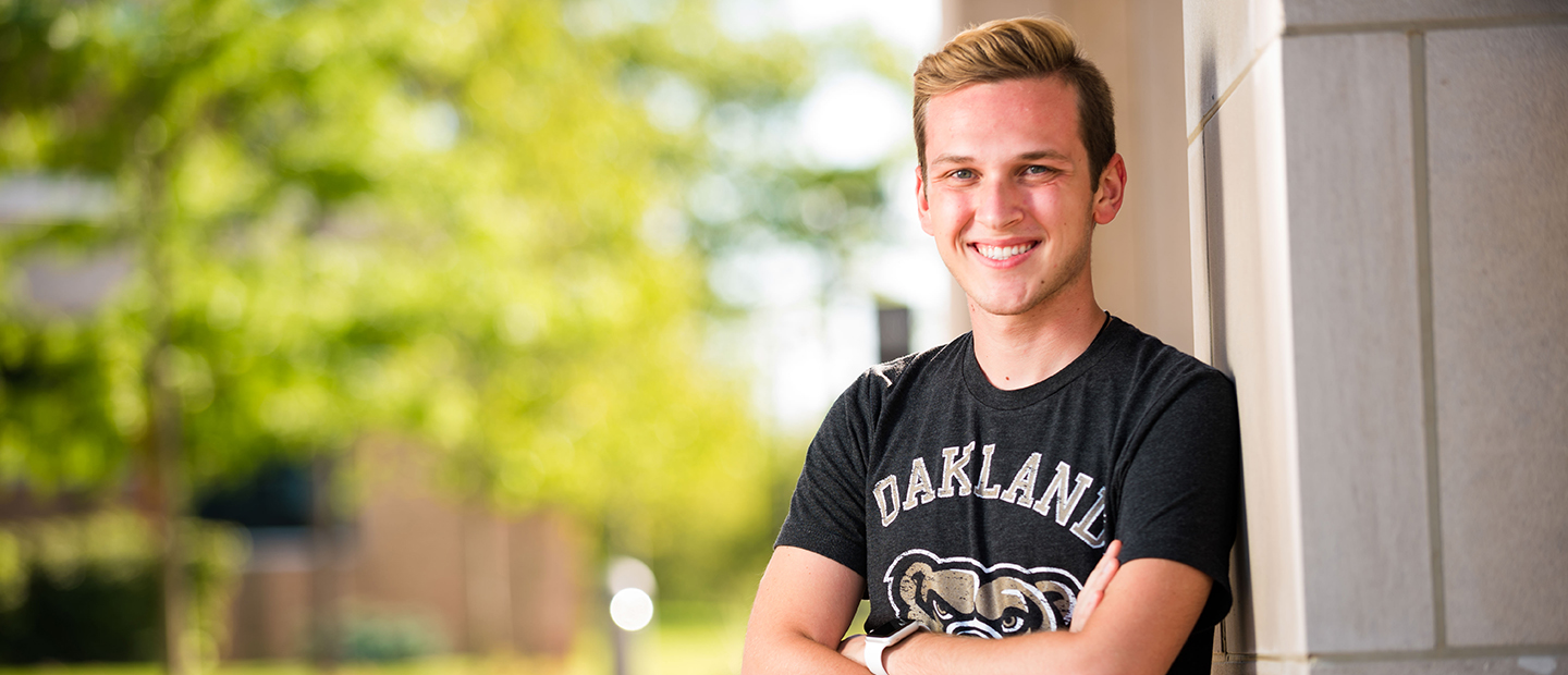 man in a black Oakland University t-shirt leaning on a wall outside