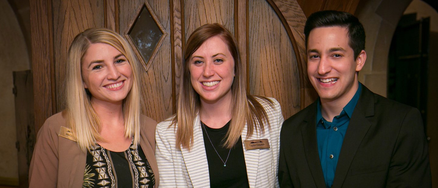 three smiling students, posing in front of a wooden door