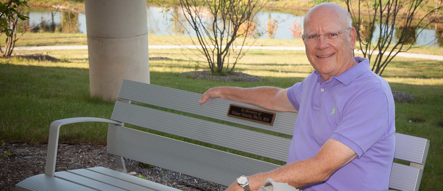 man sitting on a bench with a plaque that says "In Memory of Diane Norris, Ph.D., RN"