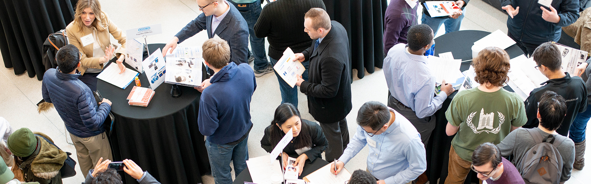 aerial photo of people standing around circular tables, looking over papers