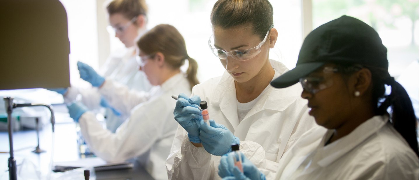 Students in white lab coats, safety glasses and blue gloves holding test tubes