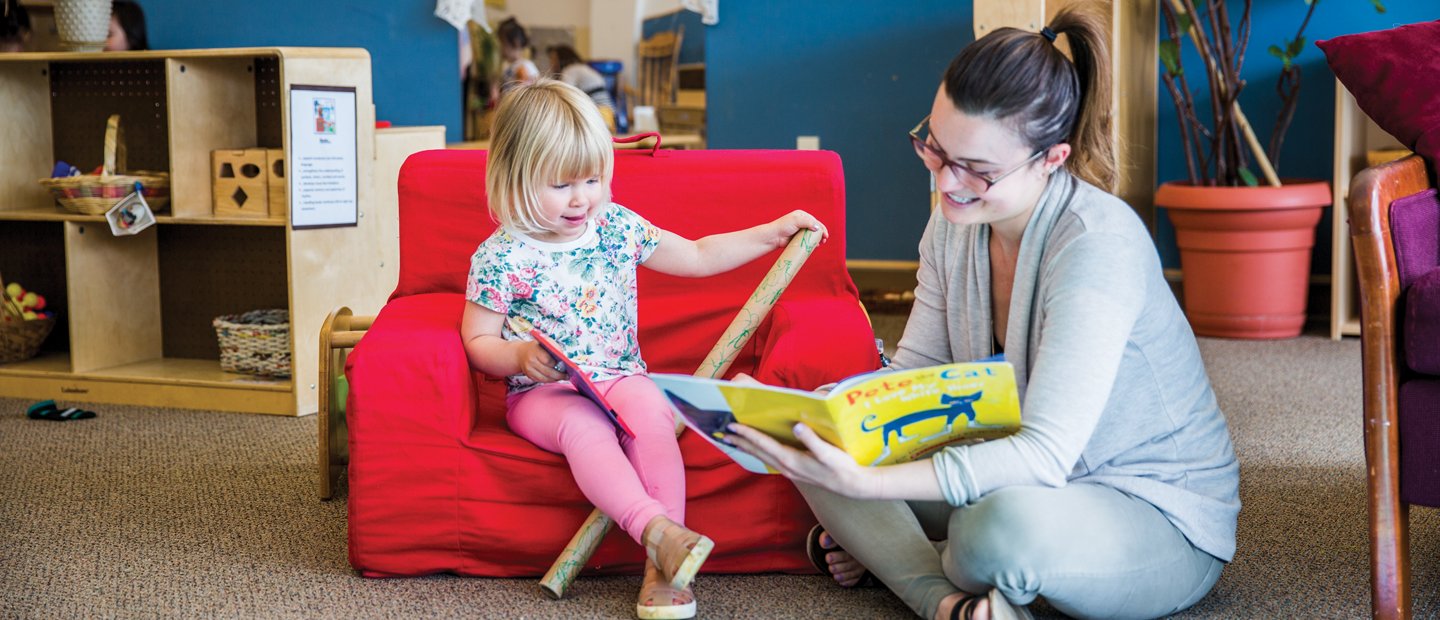 Early Childhood Education ECE Teacher reading a book to a young girl seated in a red chair in a classroom.