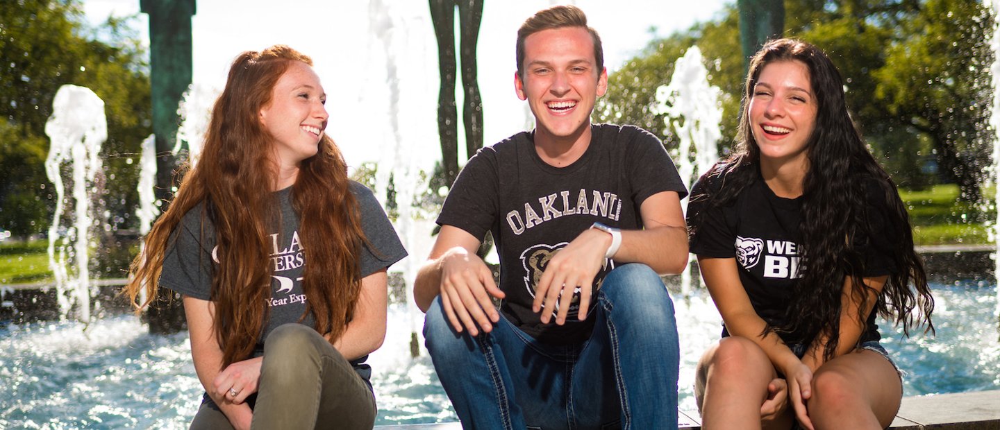 Three Oakland University students sitting on the edge of a fountain, laughing and smiling.