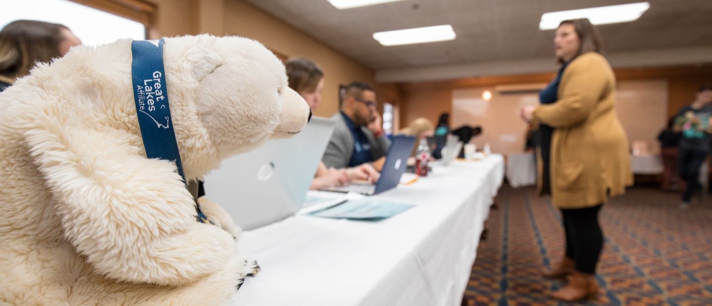Photo of polar bear stuffed animal at long table with student talking to students using computers