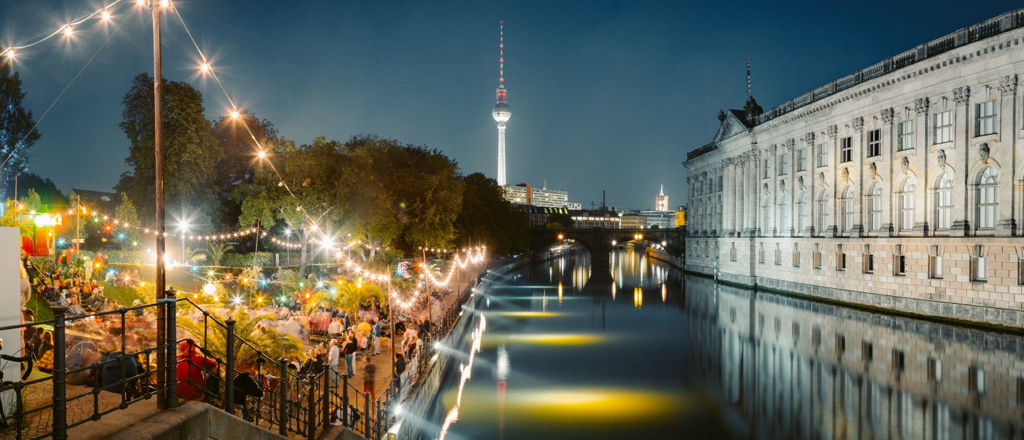 A canal in Germany with a large building on one side and a brightly lit patio on the other.