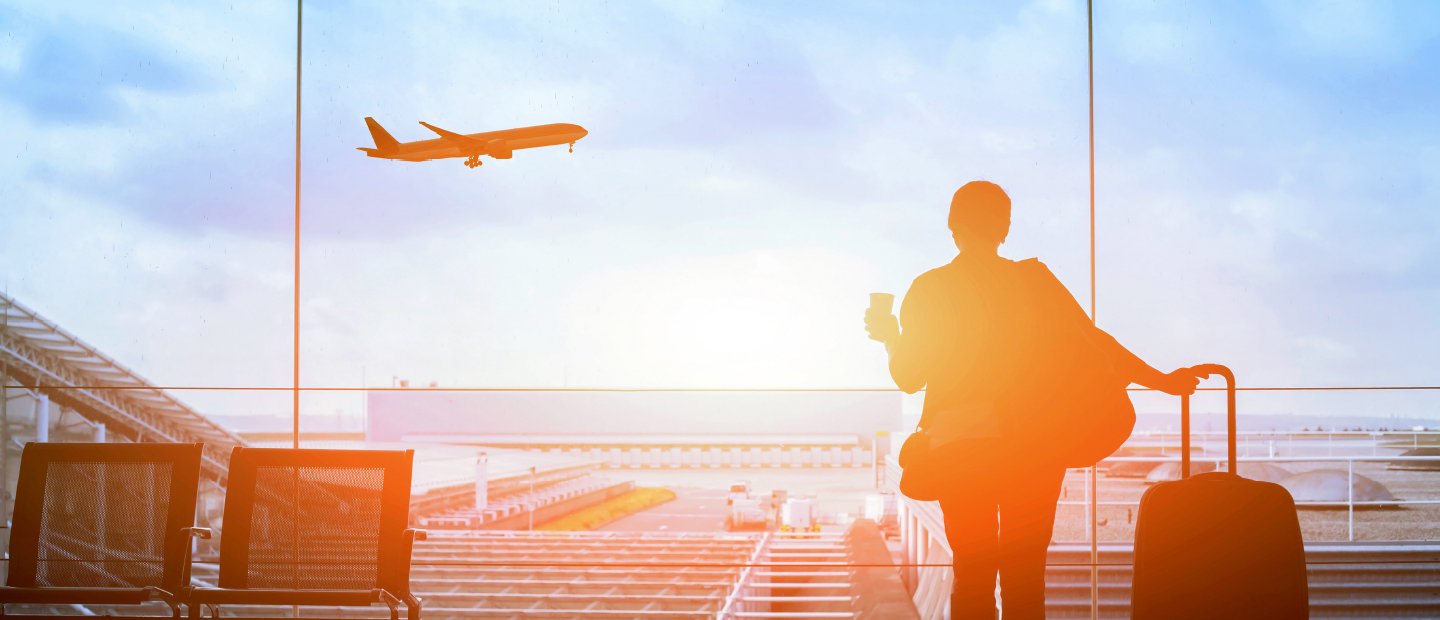 A person standing with a rolling luggage bag, watching a plane take off.