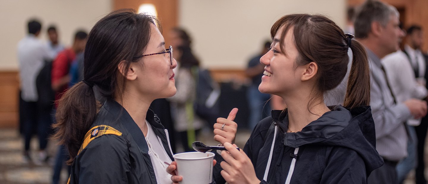 Two young women standing in a room of people, talking to each other.