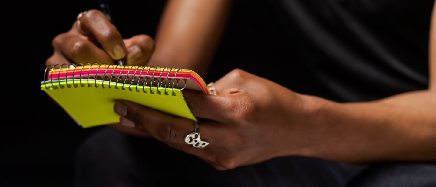 A person writing in a small, brightly colored notepad.