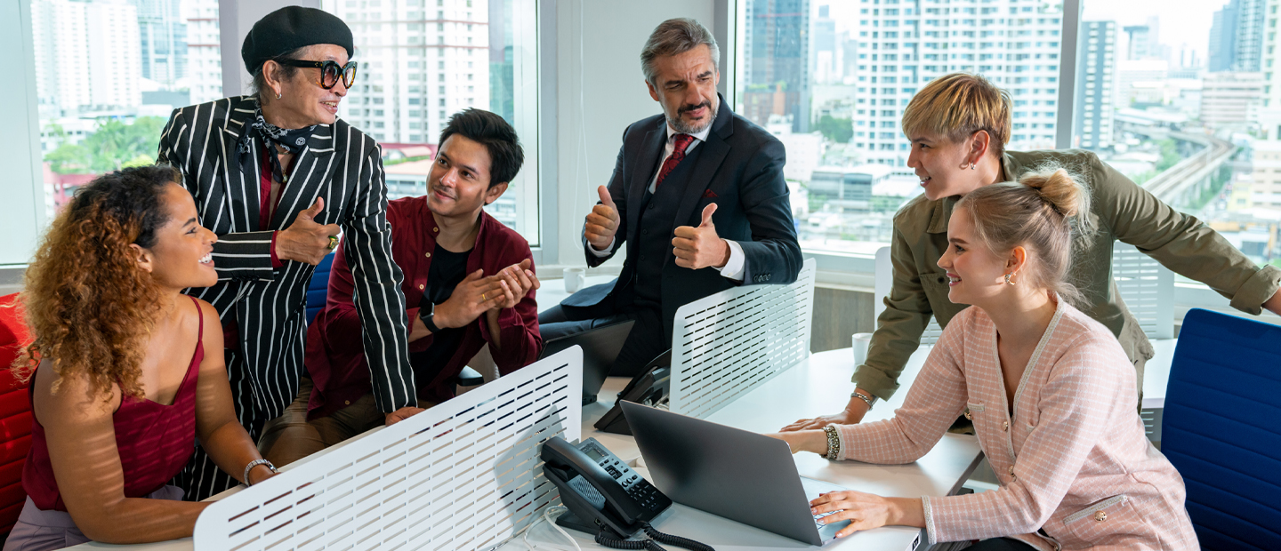 Six people sitting or standing around a desk in an office, some smiling, some giving a thumbs up signal.