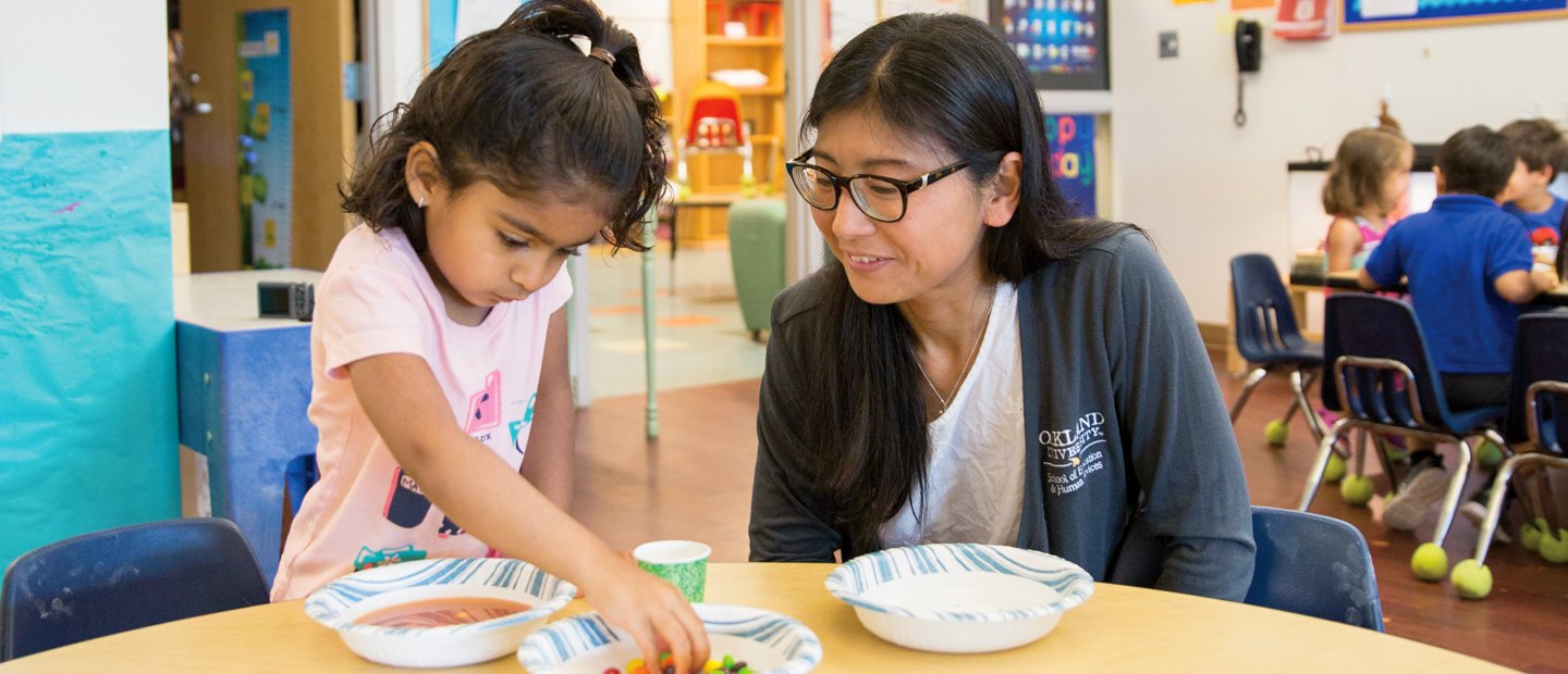Preschool A young girl sitting with a staff member, putting food into a bowl.