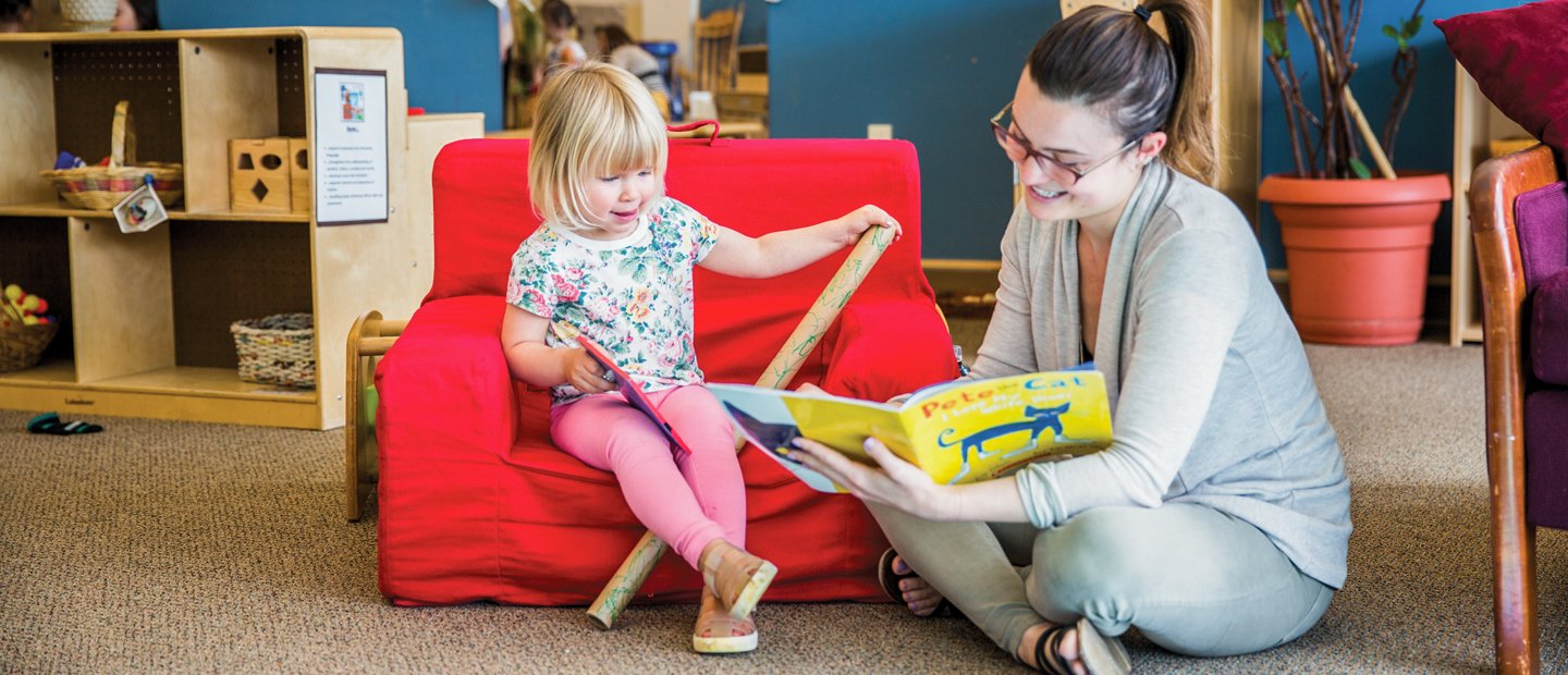 A young girl sitting in a red chair, while a staff member reads a book to her.