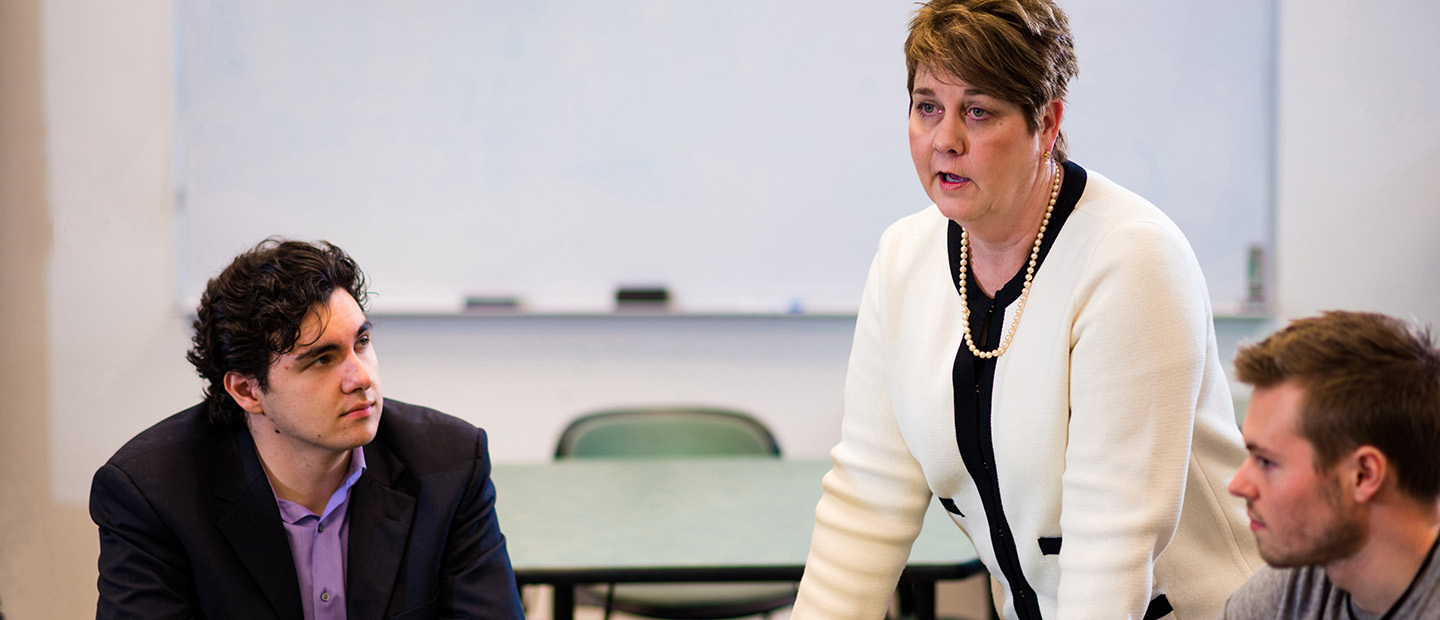 A woman standing over two adults seated at a table in a classroom.