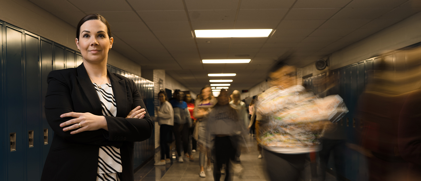 A woman standing with her arms crossed in a hallway of lockers as students walk behind her.