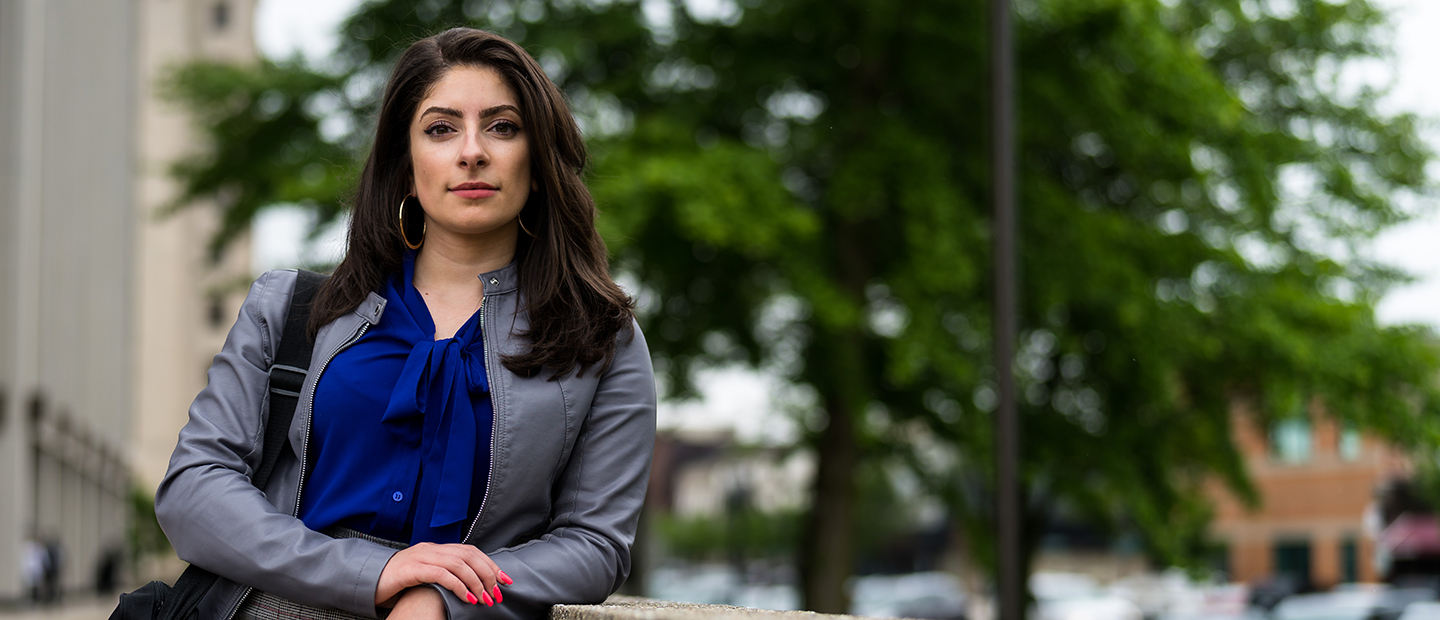 A woman standing outside, leaning on a railing, looking directly at the camera.