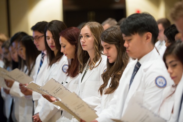 Students stand, holding programs, at the white coat ceremony.