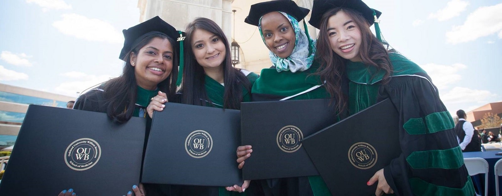 Four new OUWB graduates stand before the Elliot Tower wearing caps and gowns and holding their diplomas.