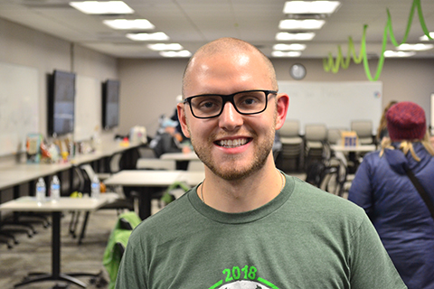 man in a green shirt, standing in a classroom, smiling at the camera