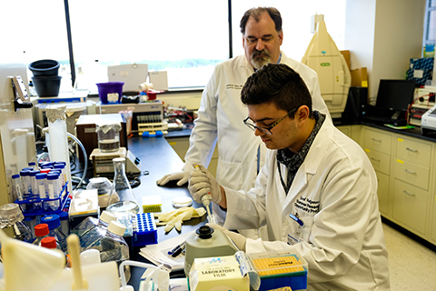 student using a medical instrument to fill a test tube in a lab with a professor watching over his shoulder