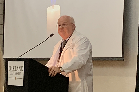 man in a white lab coat standing at a podium in front of a projector screen with a candle on it
