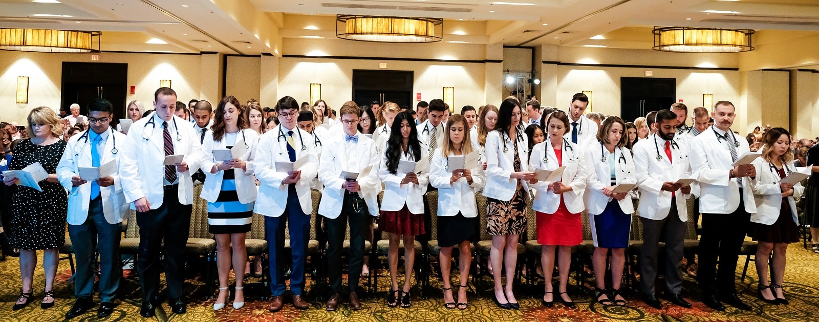 A photo of all the M1 students standing at their seats at the White Coat ceremony. They are reading from their programs.