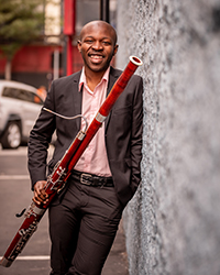 Jaquain Sloan holding a bassoon, leaning against a concrete wall outside.