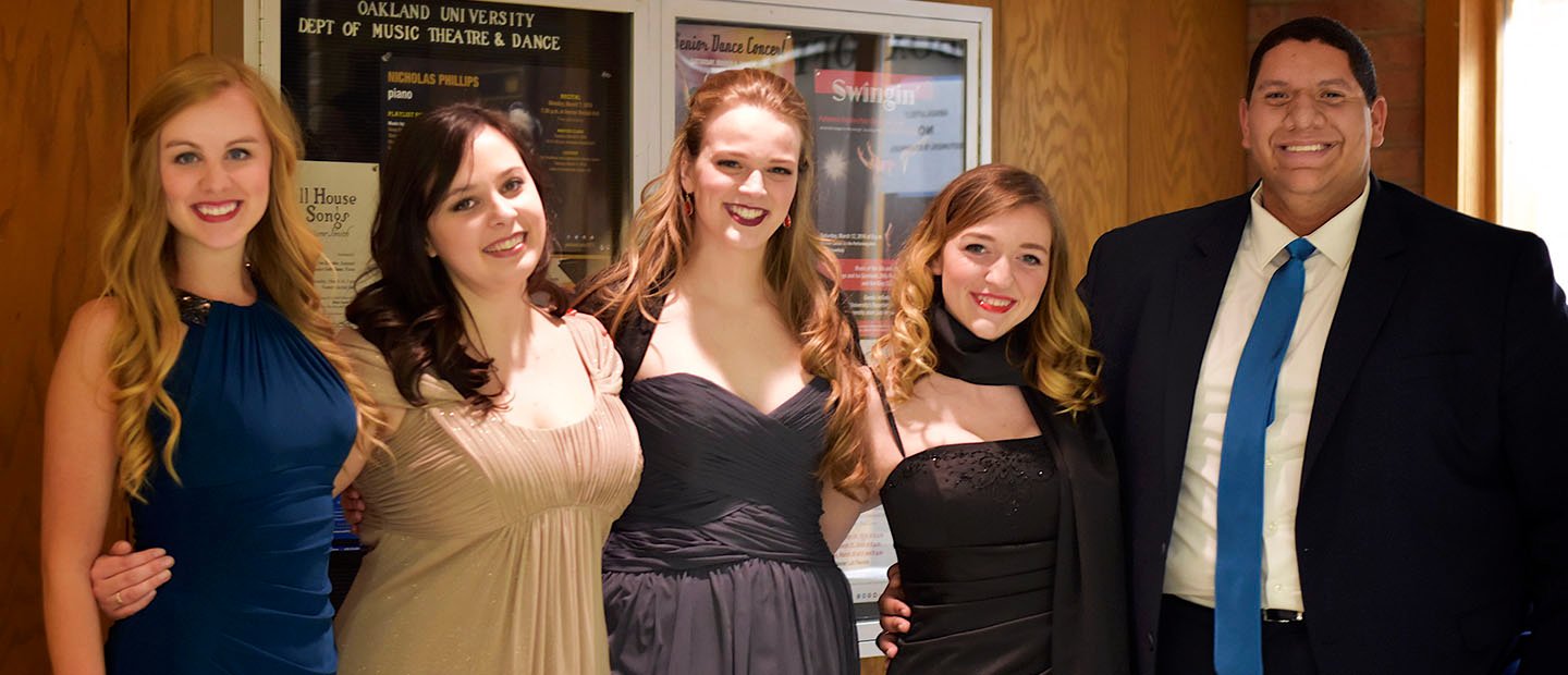 five people in evening attire, standing in a row with arms around each other, smiling at the camera