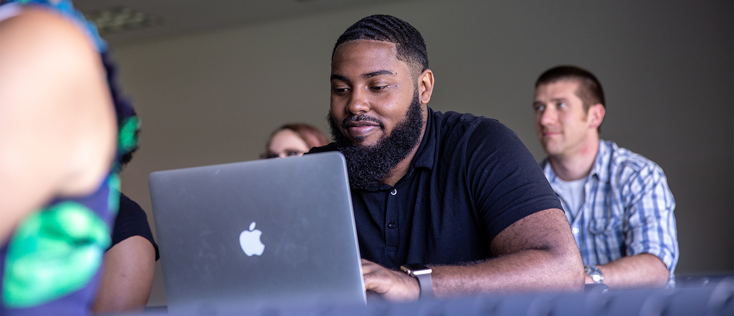 Image of a student wearing a black shirt in a classroom looking at his computer. 