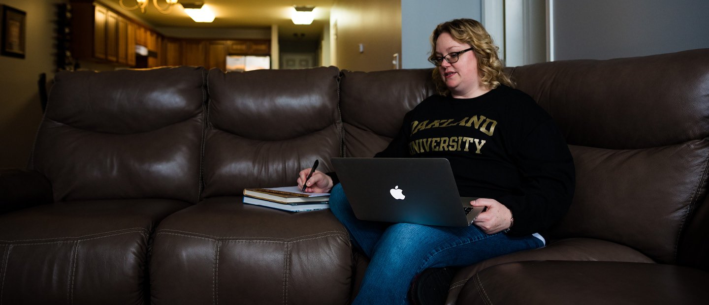 woman seated on a large leather couch with a laptop in her lap, writing in a notebook