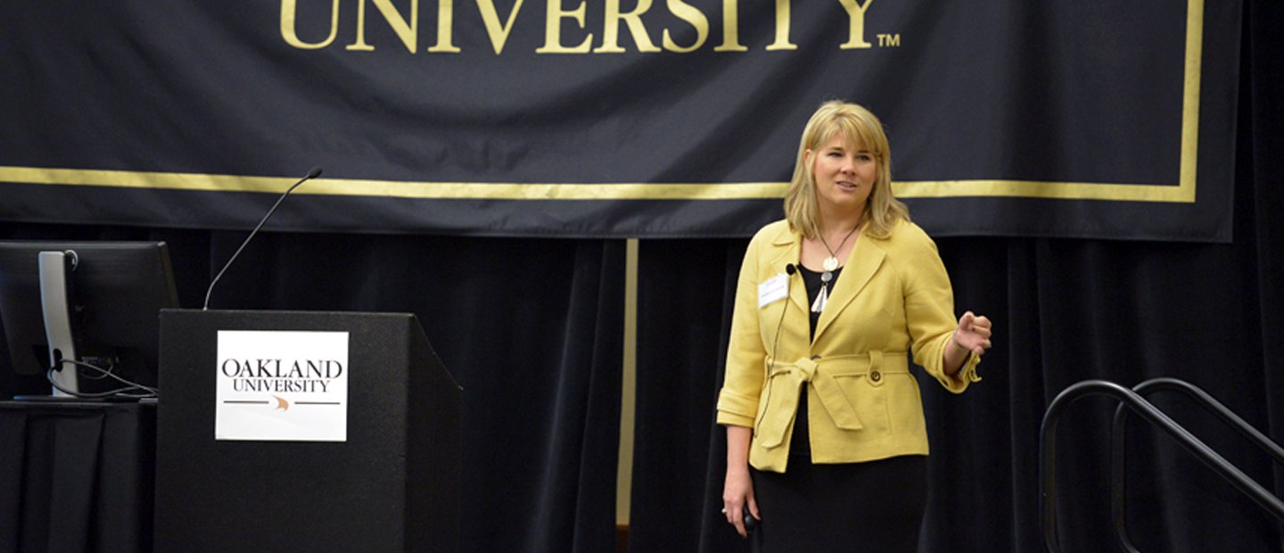 woman speaking on a stage with an Oakland University banner behind her