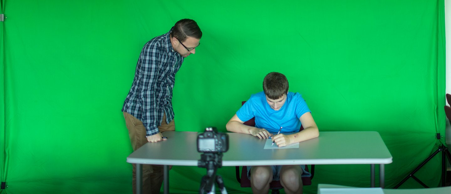 A boy seated at a table in front of a green screen, writing while a teacher looks over his shoulder.
