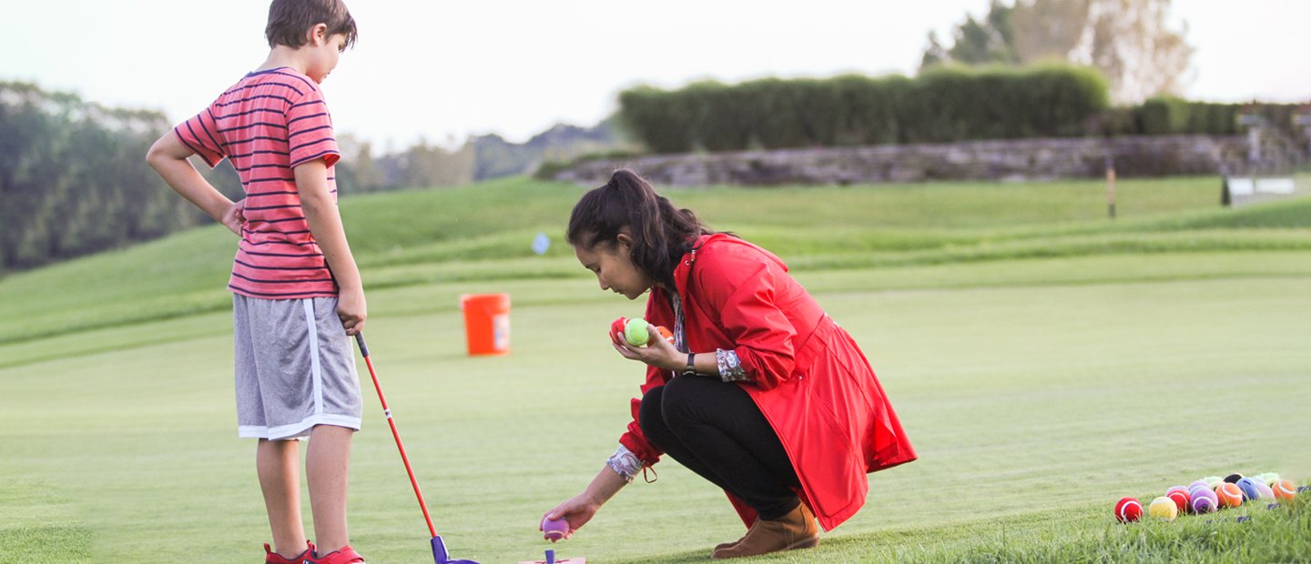 A woman putting a ball on a golf tee next to a young boy holding a club at a golf course.