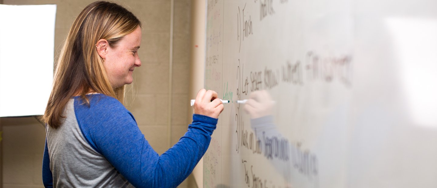 A young woman writing on a white board and smiling.