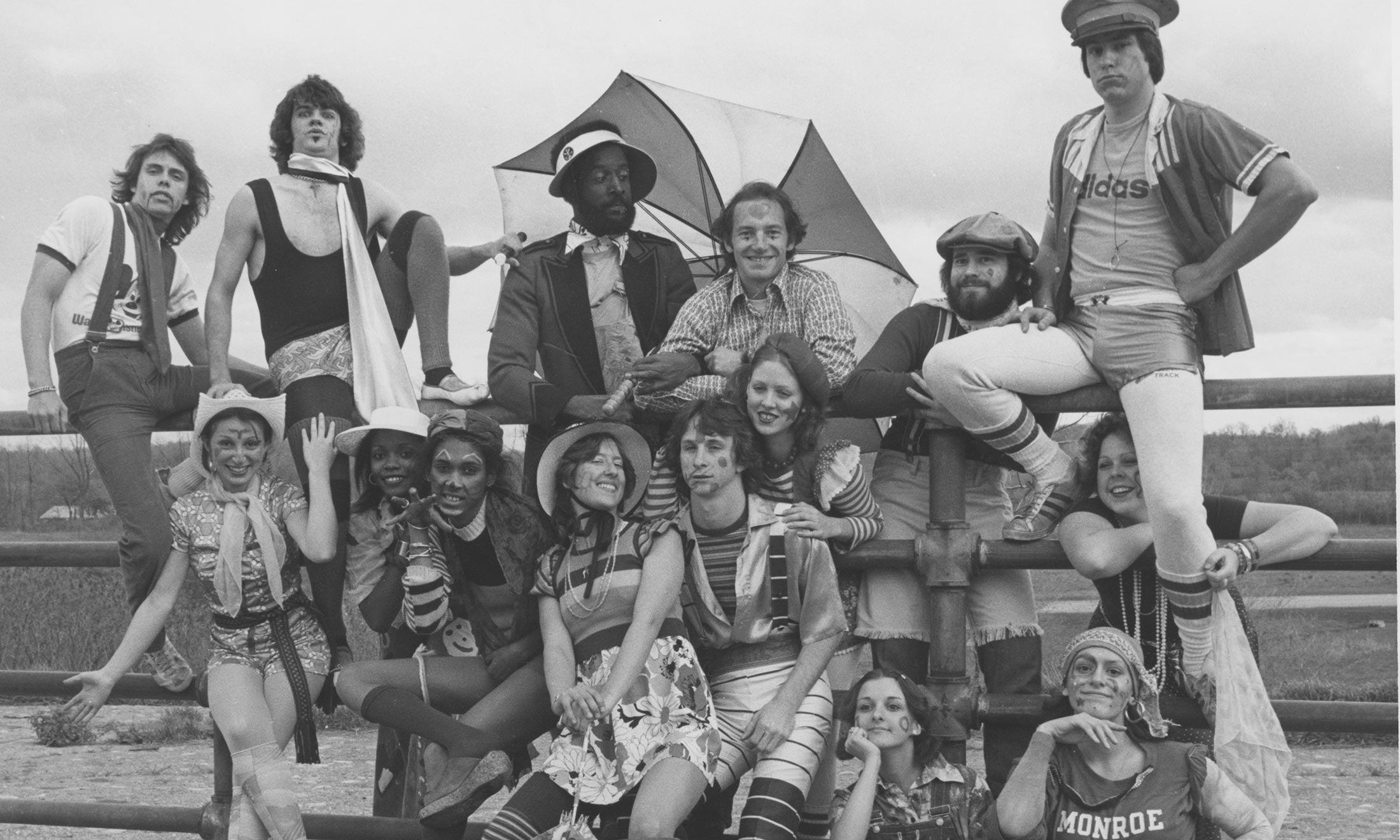 Oakland University drama students dressed in costumes sit on a gate for a photo outside in 1975