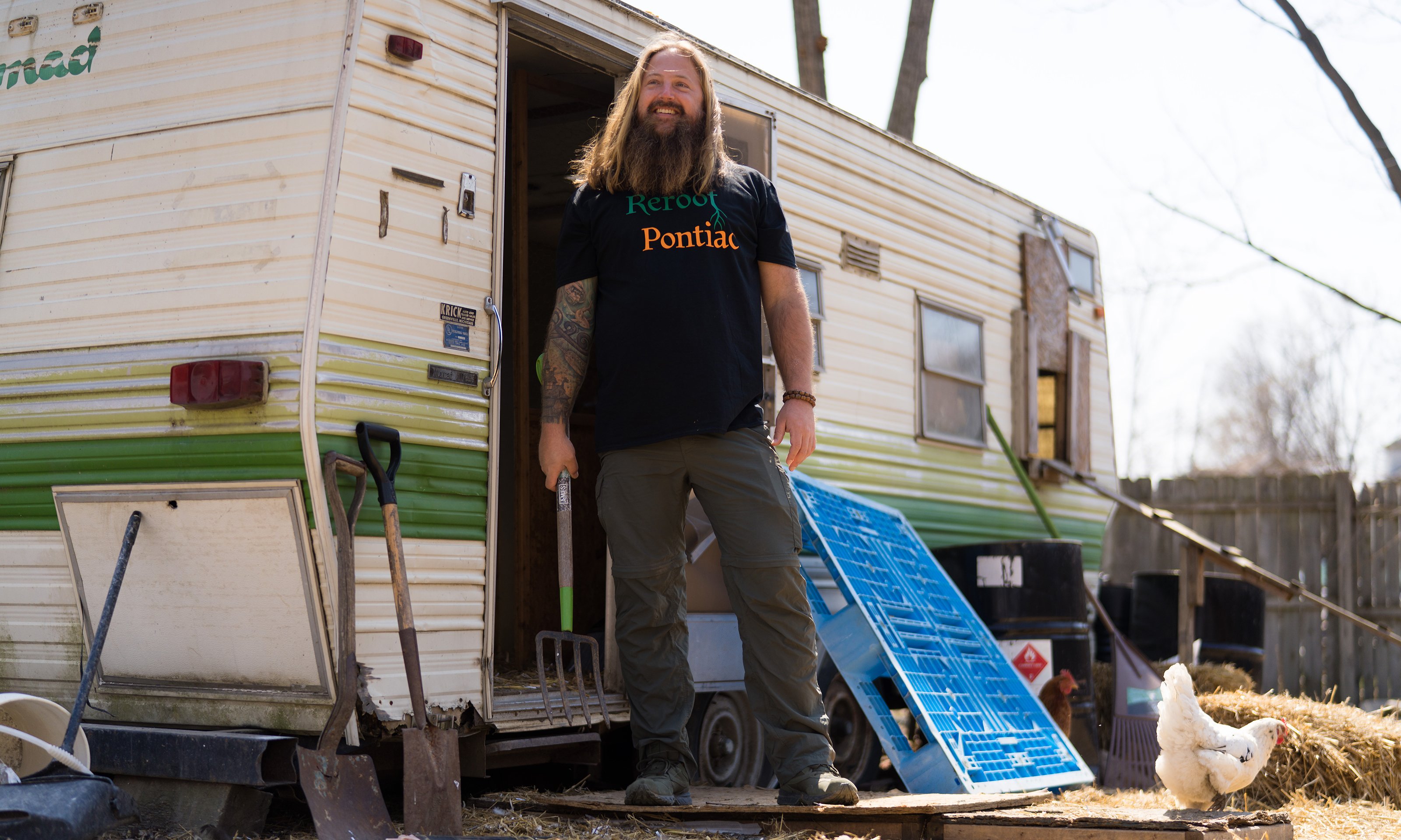 A man holding a pitchfork standing outside of a mobile home.