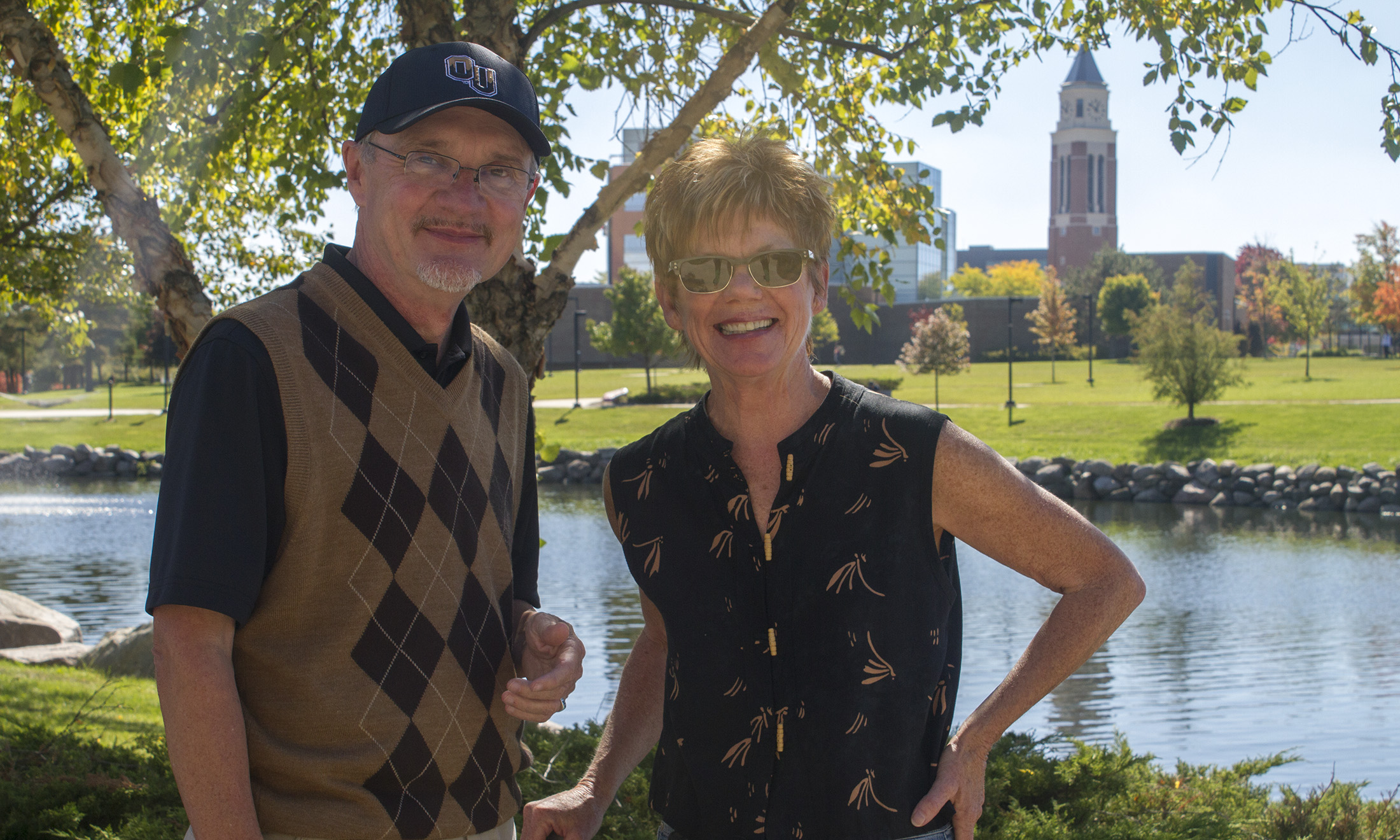 Photo os Garry Gilbert and Holly Gilbert standing next to lake with Oakland University campus in background