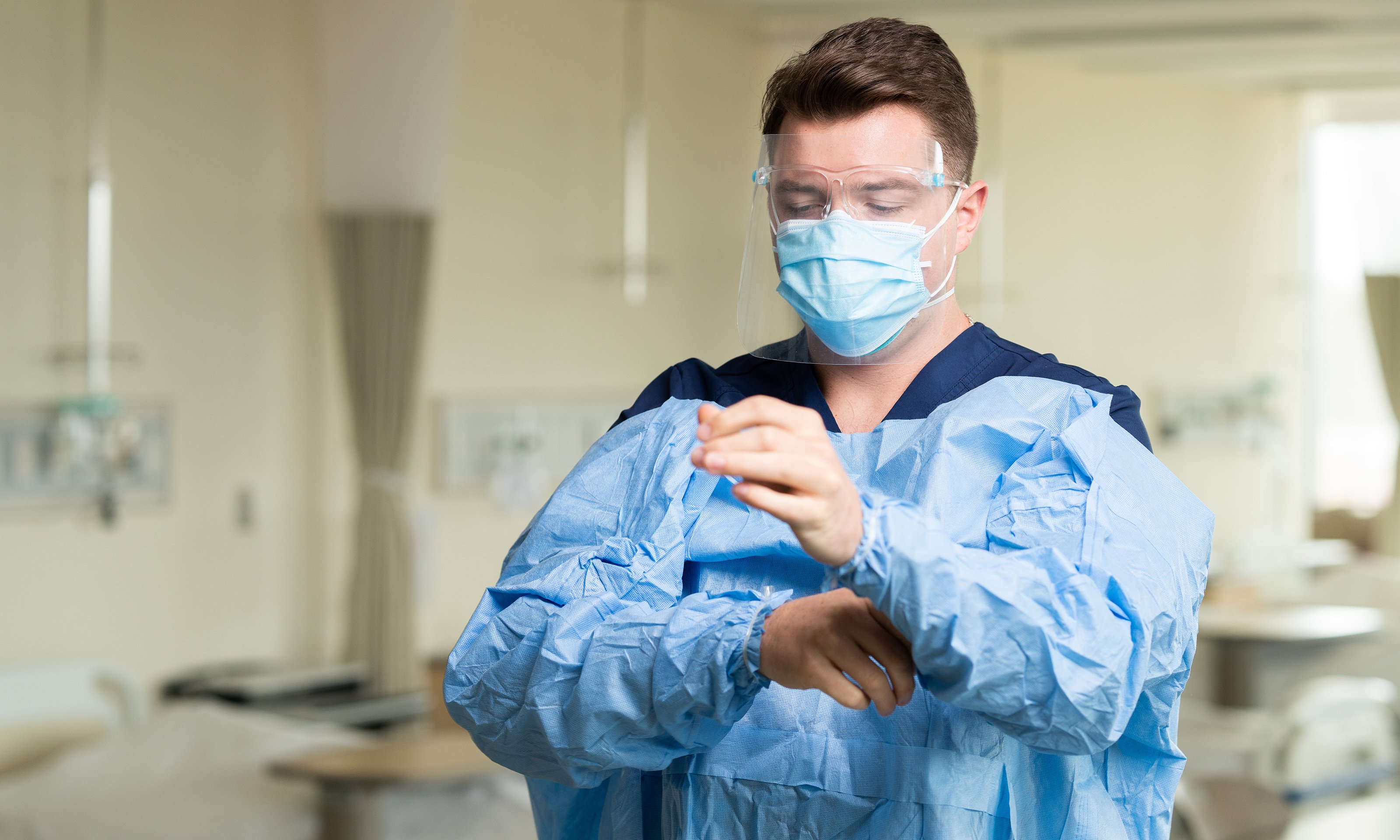 A man wearing a N95, surgical mask and face shield donning a surgical gown.