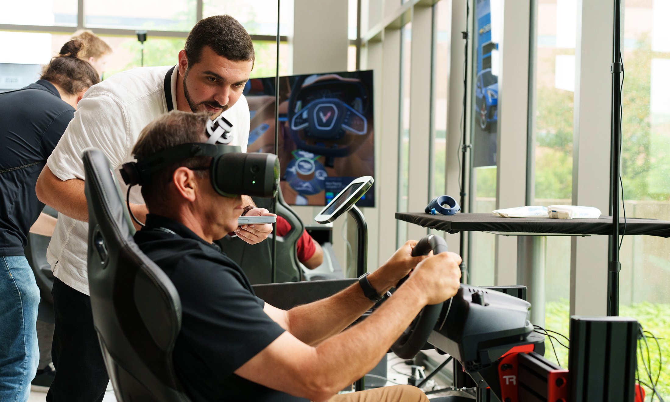 A person watching someone wearing a VR headset and using a steering wheel.