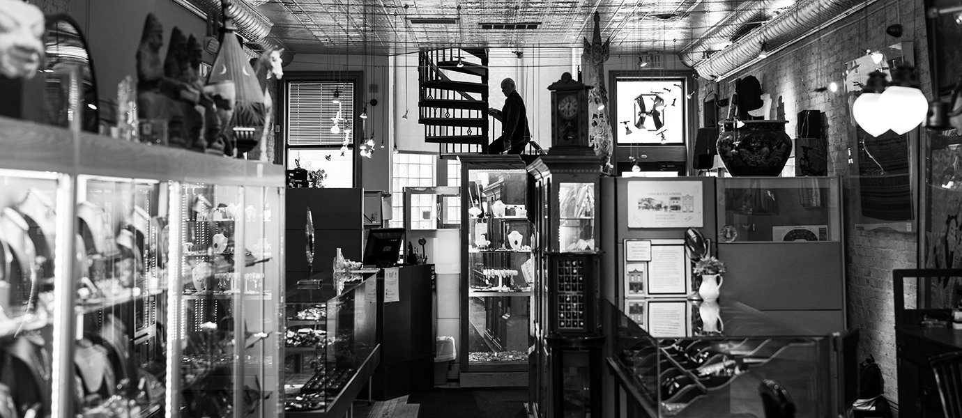 A black and white view down a jewelry store with a silhouette of a man walking up a winding staircase 