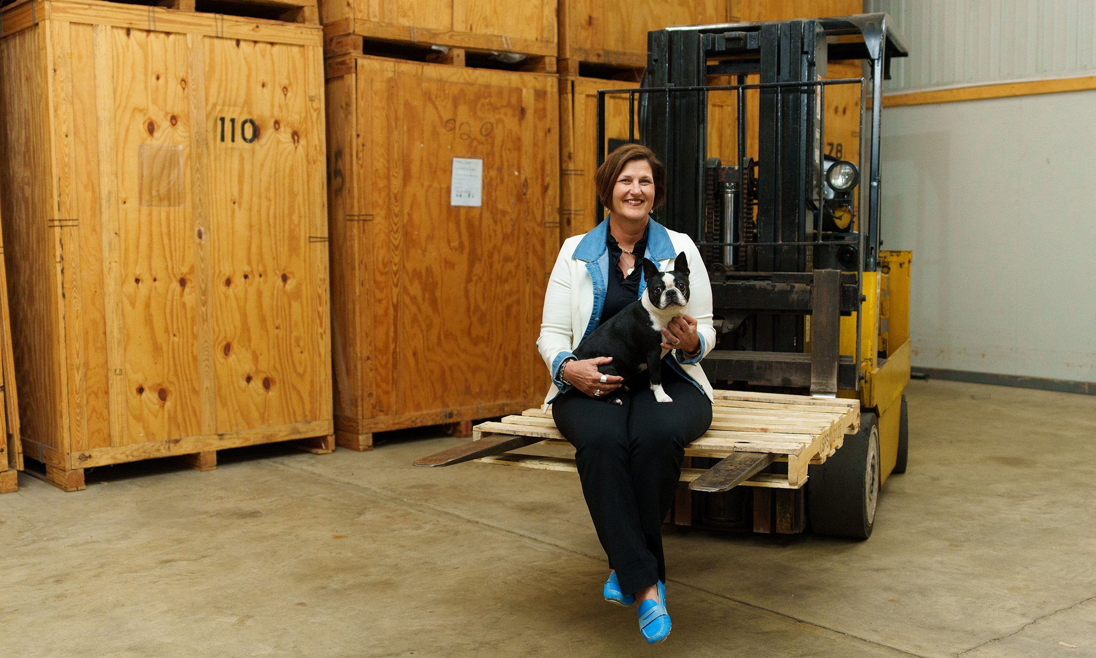 A woman holding her dog, sitting on a pallet that's on a forklift
