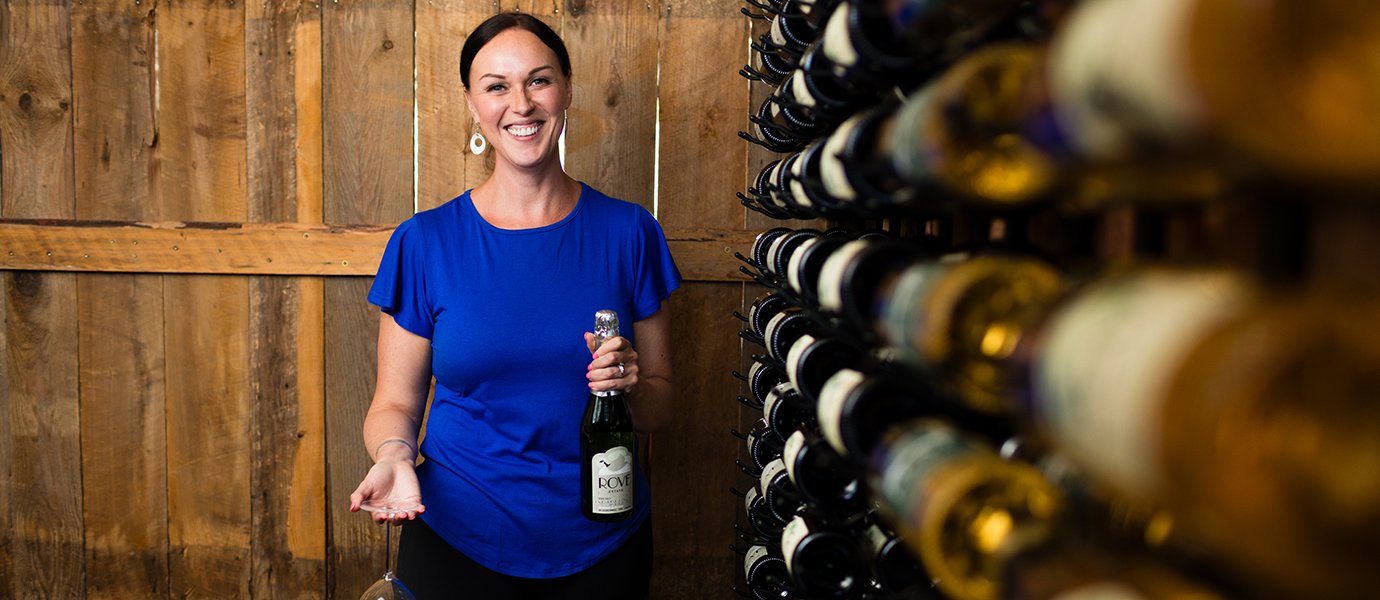 A woman smiling with a wine bottle next to a wine bottle rack