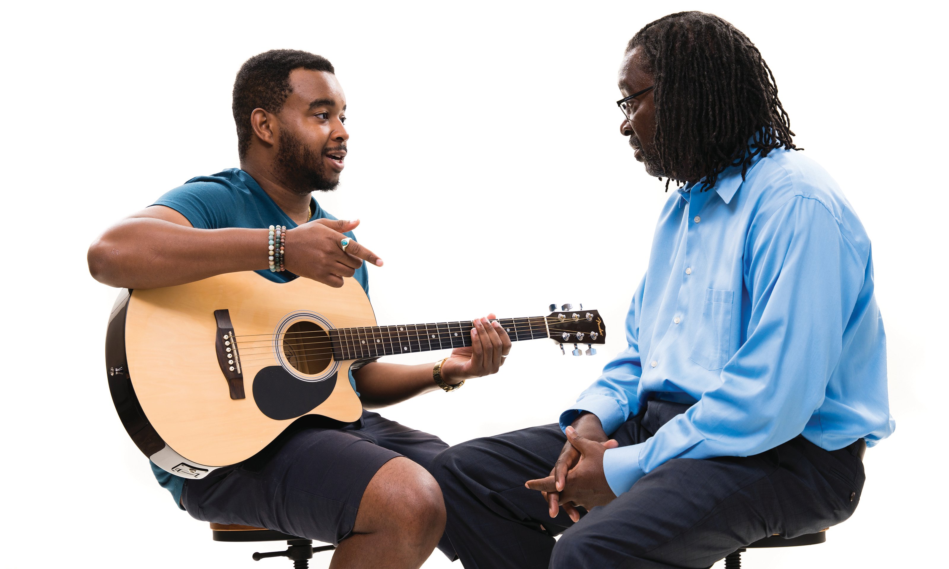 A man sitting and holding a guitar talking to another man.