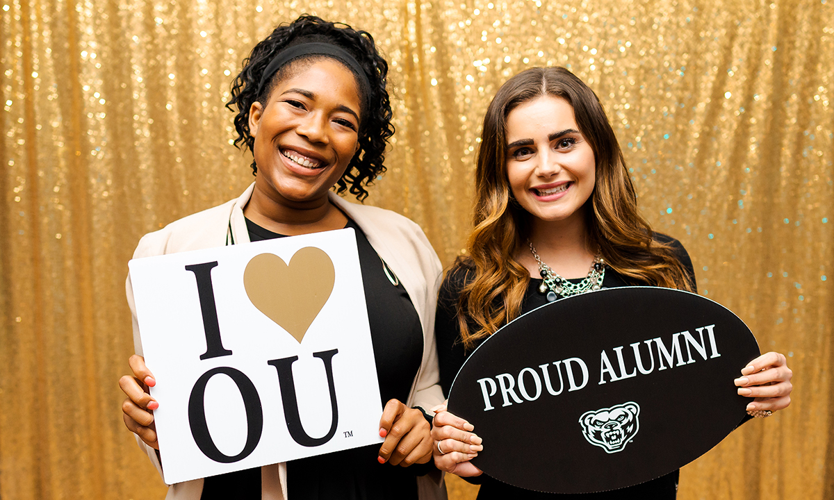 Two women standing together holding signs with text "I heart OU" and "Proud Alumni"