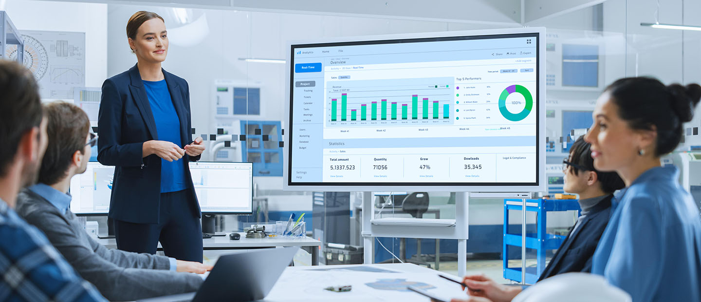 A woman presenting in front of a monitor displaying graphs.