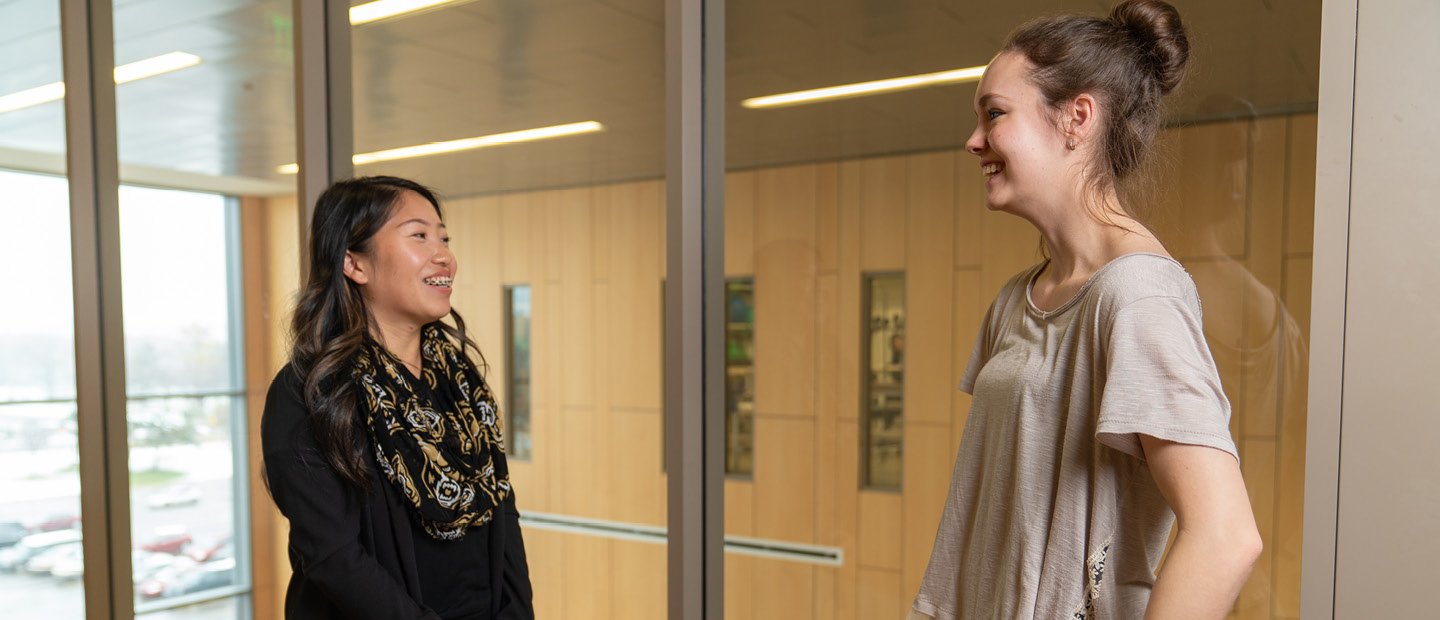 Two women standing in a room with glass walls, smiling and talking.