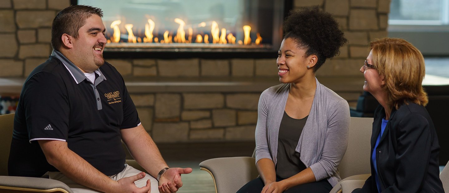 Two women and a man seated in front of a fire place, smiling and talking.