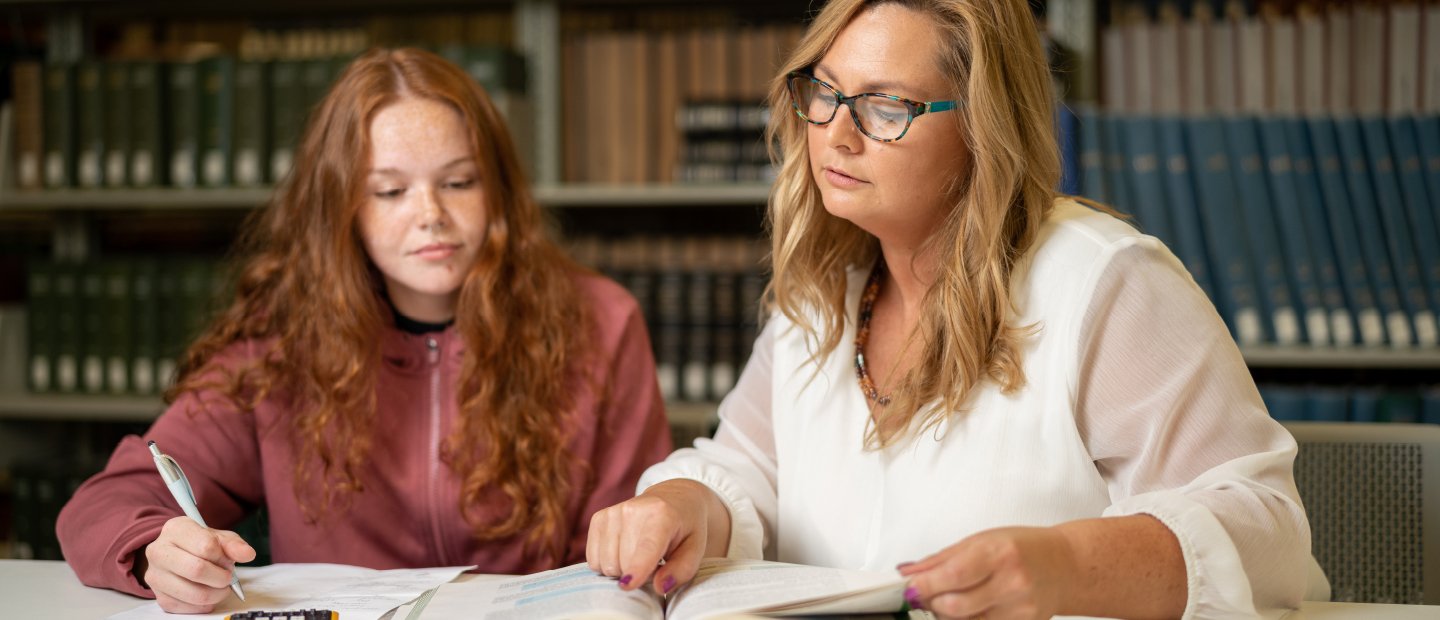 Two women seated at a table in a library, reading a book together.