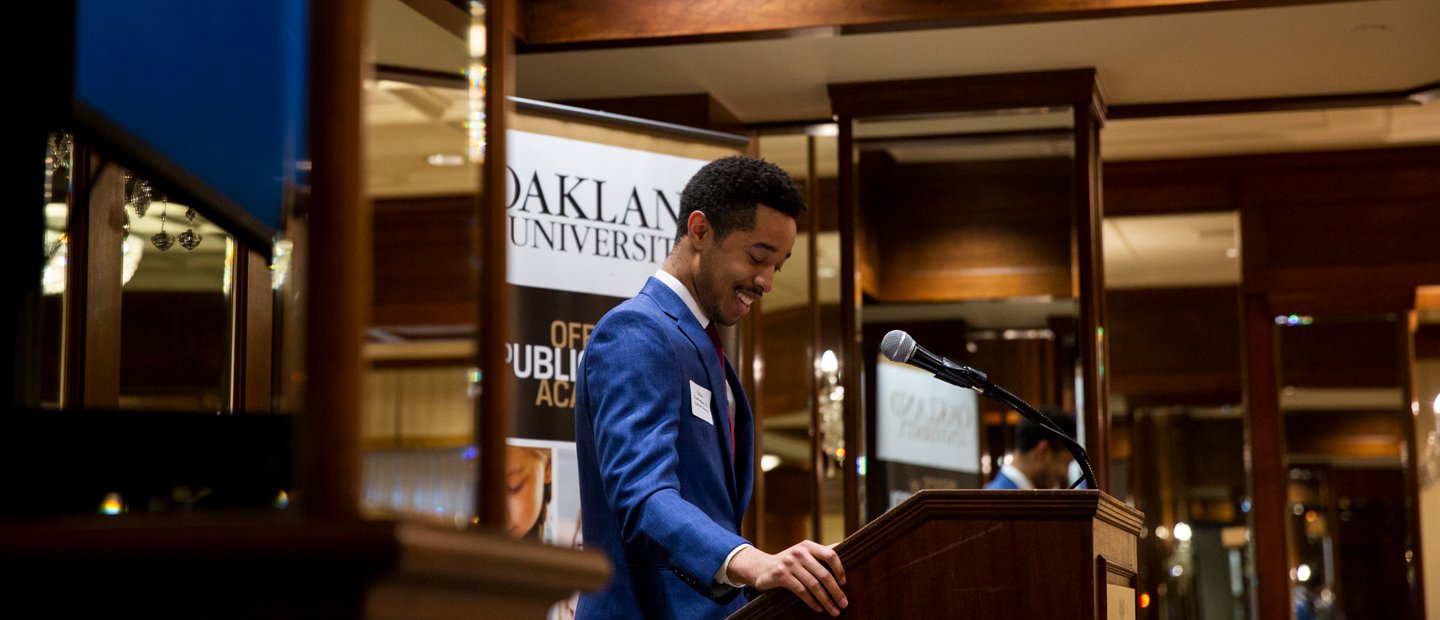 School Resources A man standing at a podium with a microphone, in front of an Oakland University banner.