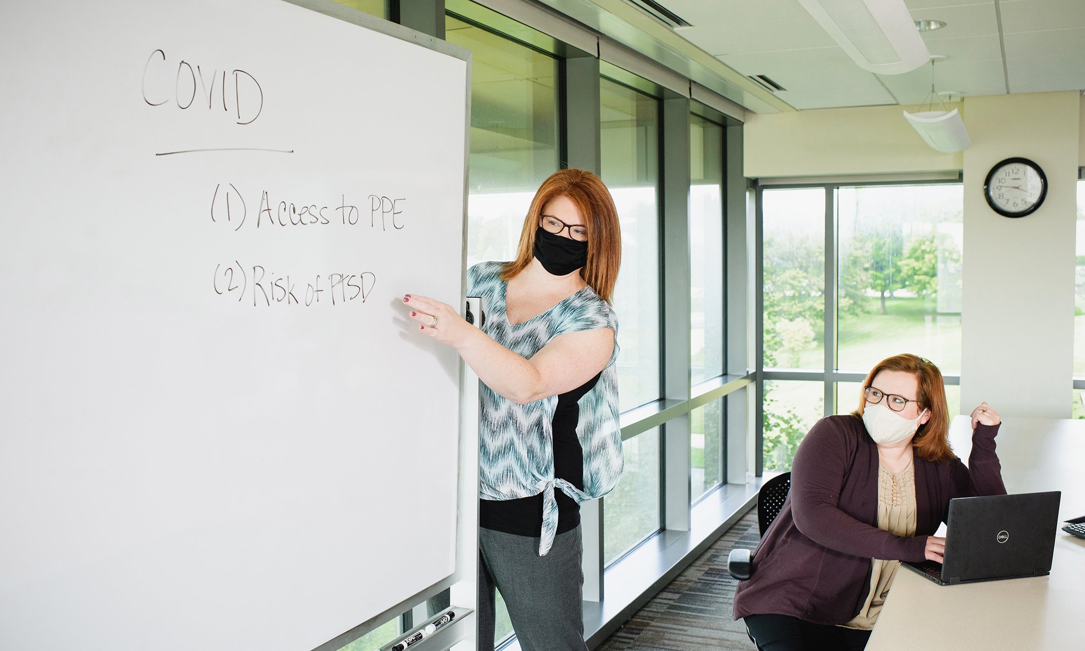 One woman writing on a white board, the other sitting. Both are wearing face masks.